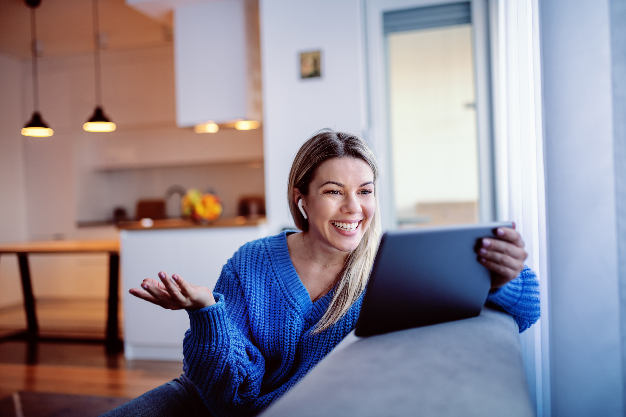 Gorgeous smiling caucasian young blonde in blue sweater and in jeans sitting on sofa and using tablet for video call. Living room interior.