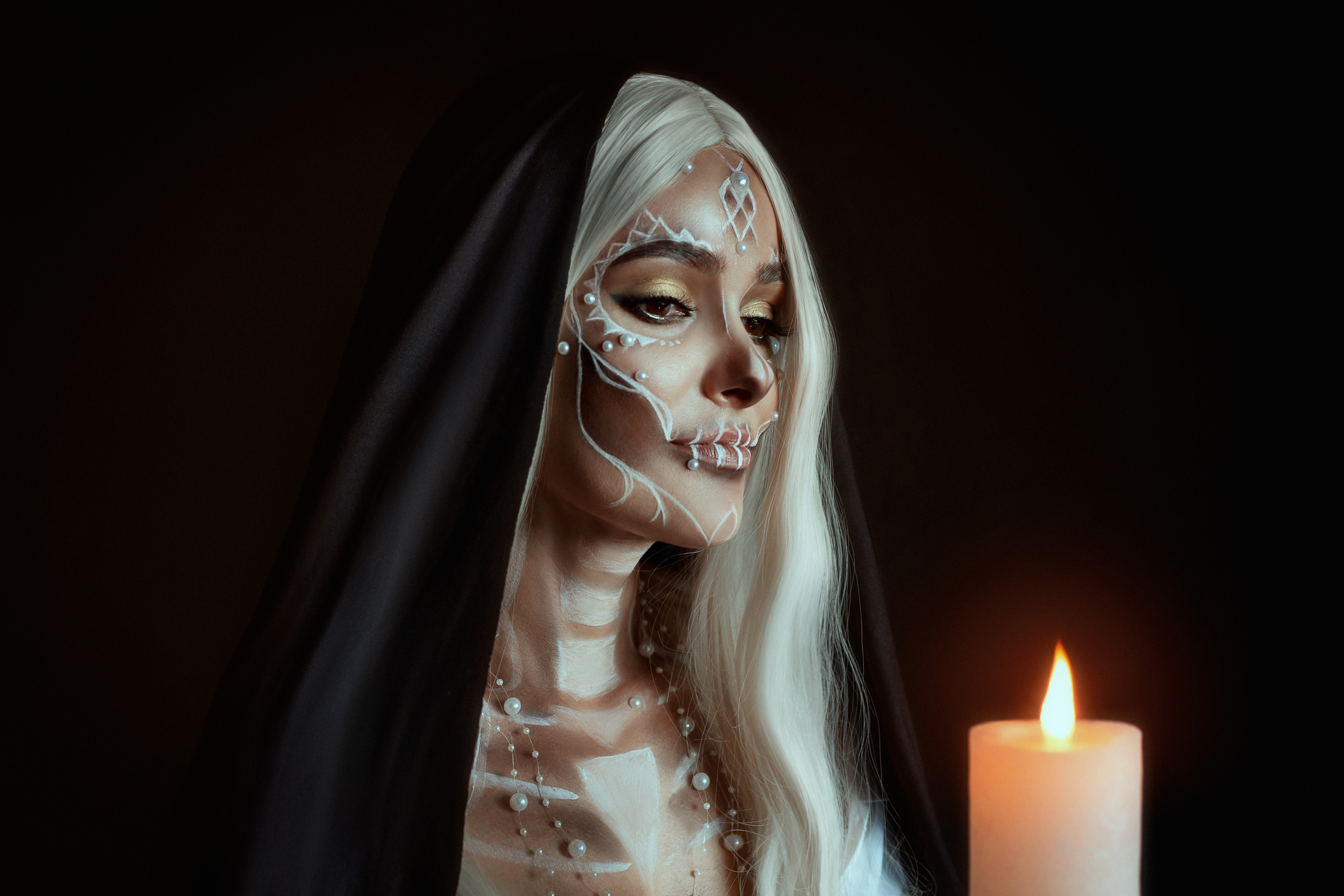 A woman in the image of an attractive mysterious witch with white ashy hair holds a burning candle. Creative makeup for Halloween party. Black poppy and dark backdrop. Celebrating Dia de los Muertos