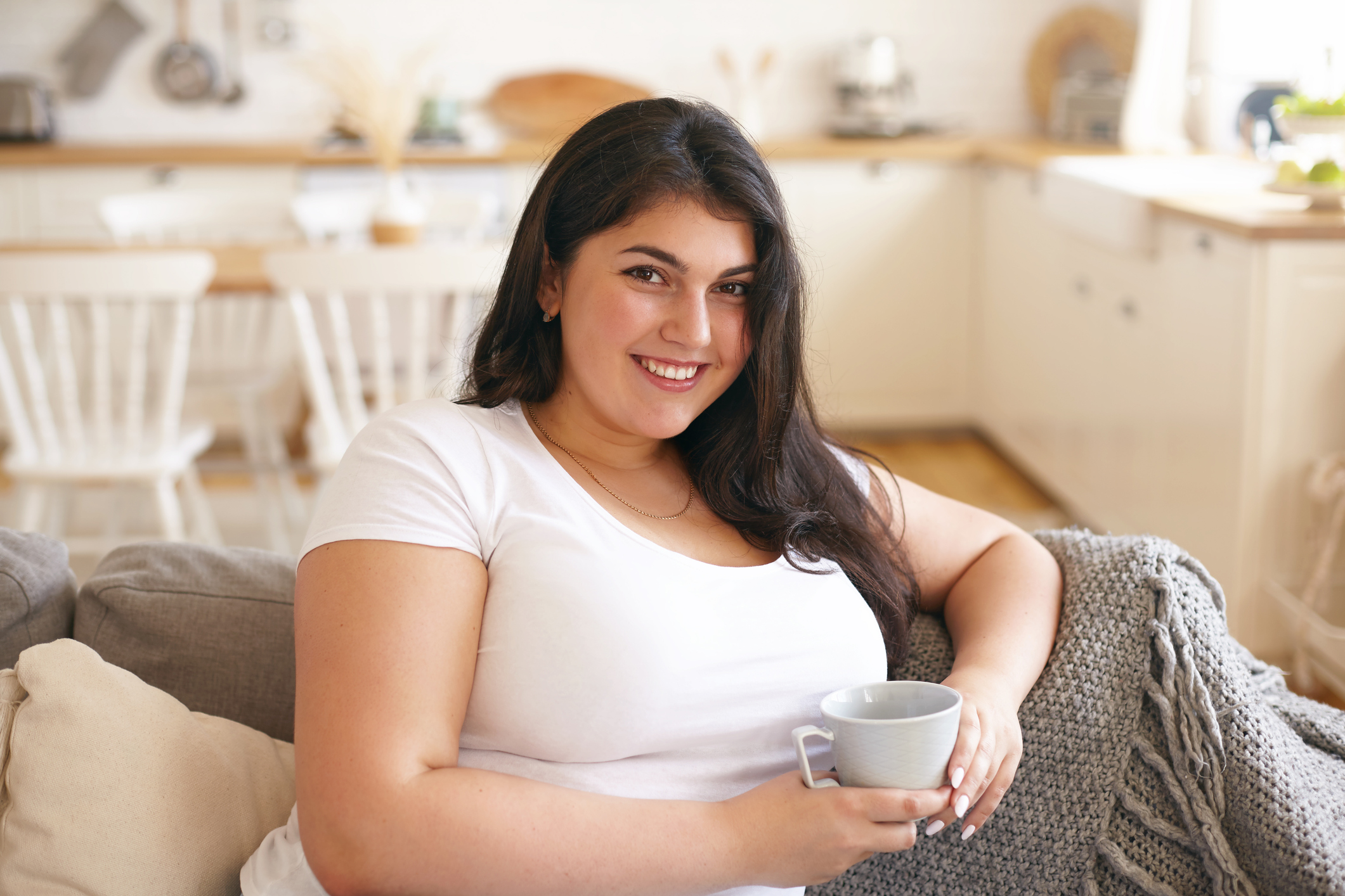 Rest, leisure and relaxation concept. Portrait of beautiful young brunette woman with curvy body and charming smile enjoying nice time being on her own, sitting on couch with cup of coffee