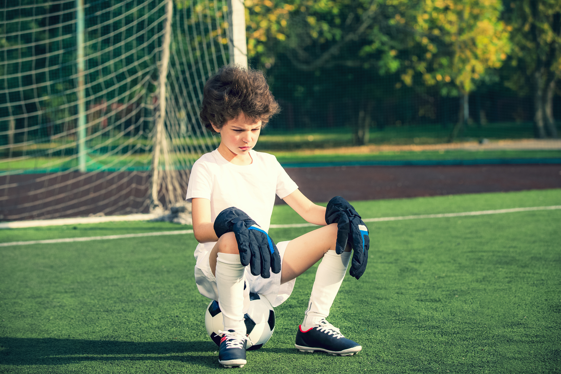 Summer soccer tournament.emotions and joy of the game. Young goals sitting on a ball. Boy goalkeeper in football sportswear on stadium with ball. Sport concept.