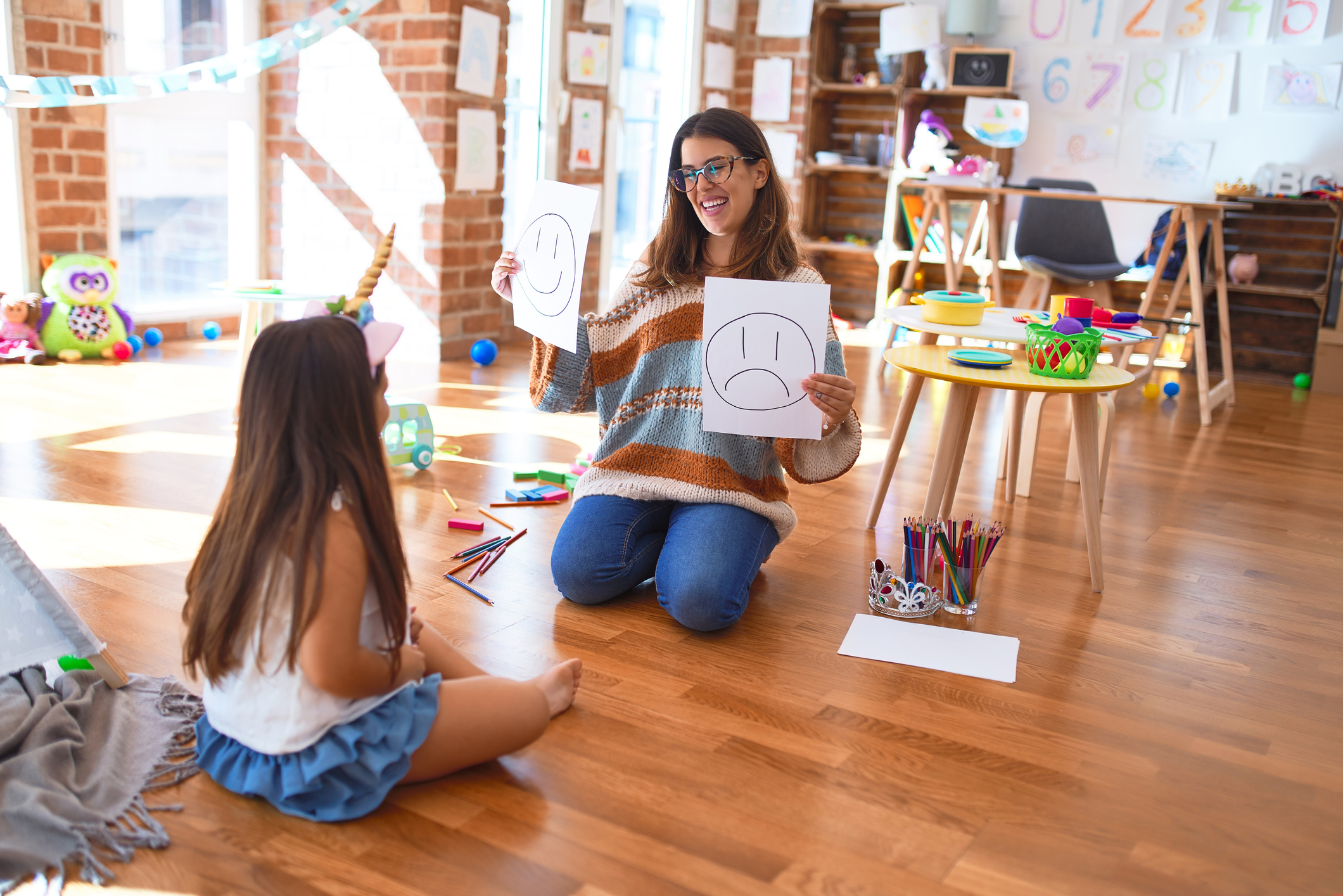 Beautiful pshychologist and toddler doing therapy with emotions emoji around lots of toys at kindergarten