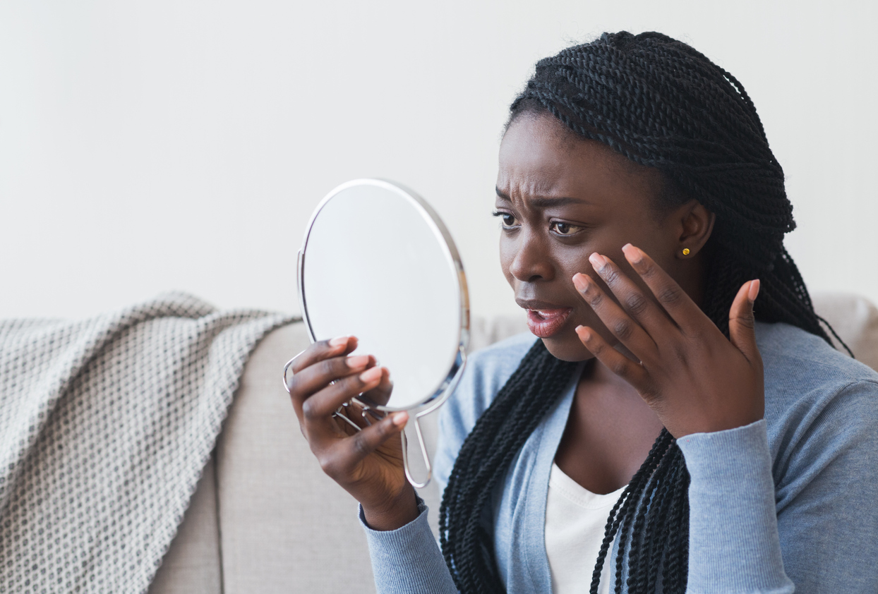 Disappointed afro woman looking to mirror and touching her skin