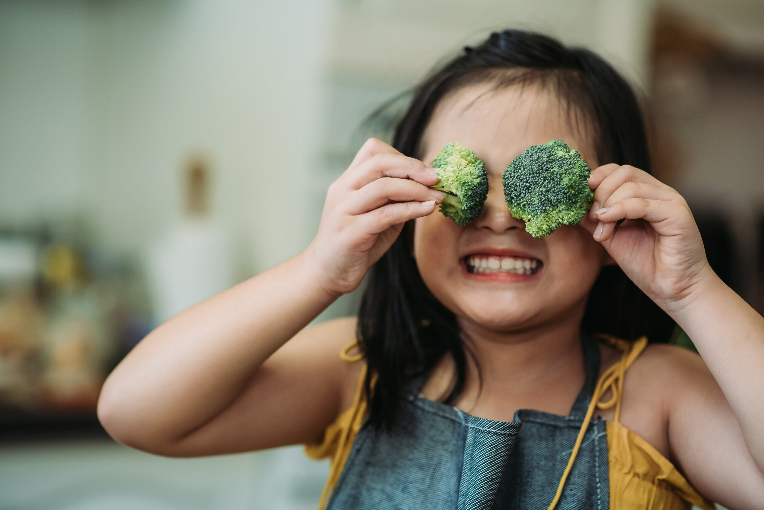 Asian chinese female child act cute with hand holding broccoli putting in front of her eyes with smiling face at kitchen