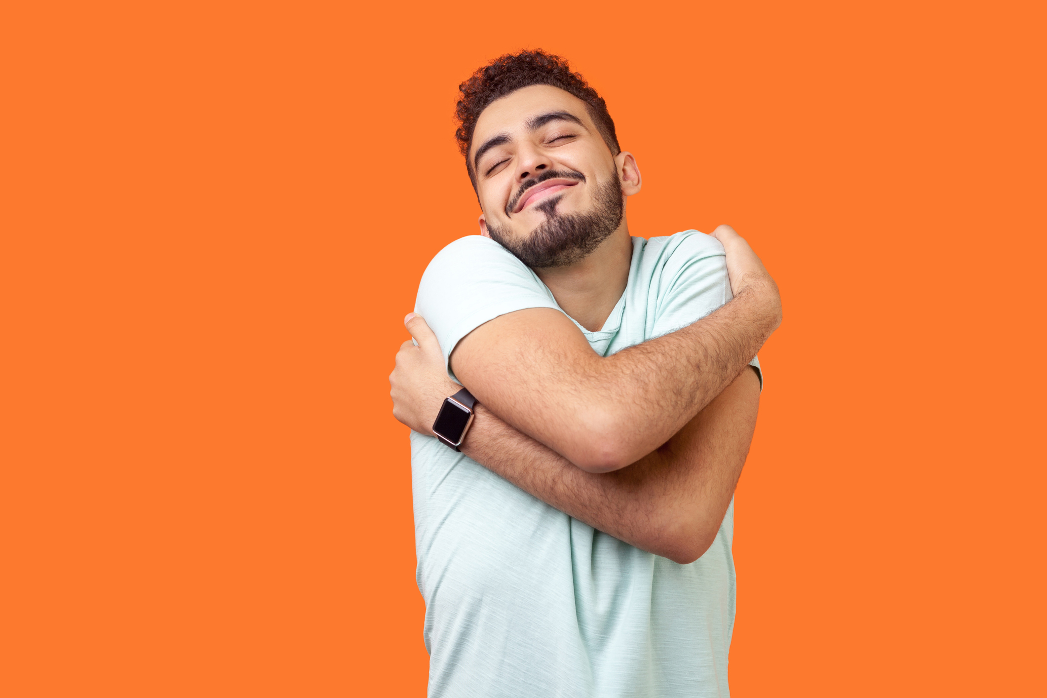 I love myself! Portrait of egoistic brunette embracing himself and smiling form pleasure. indoor studio shot isolated on orange background