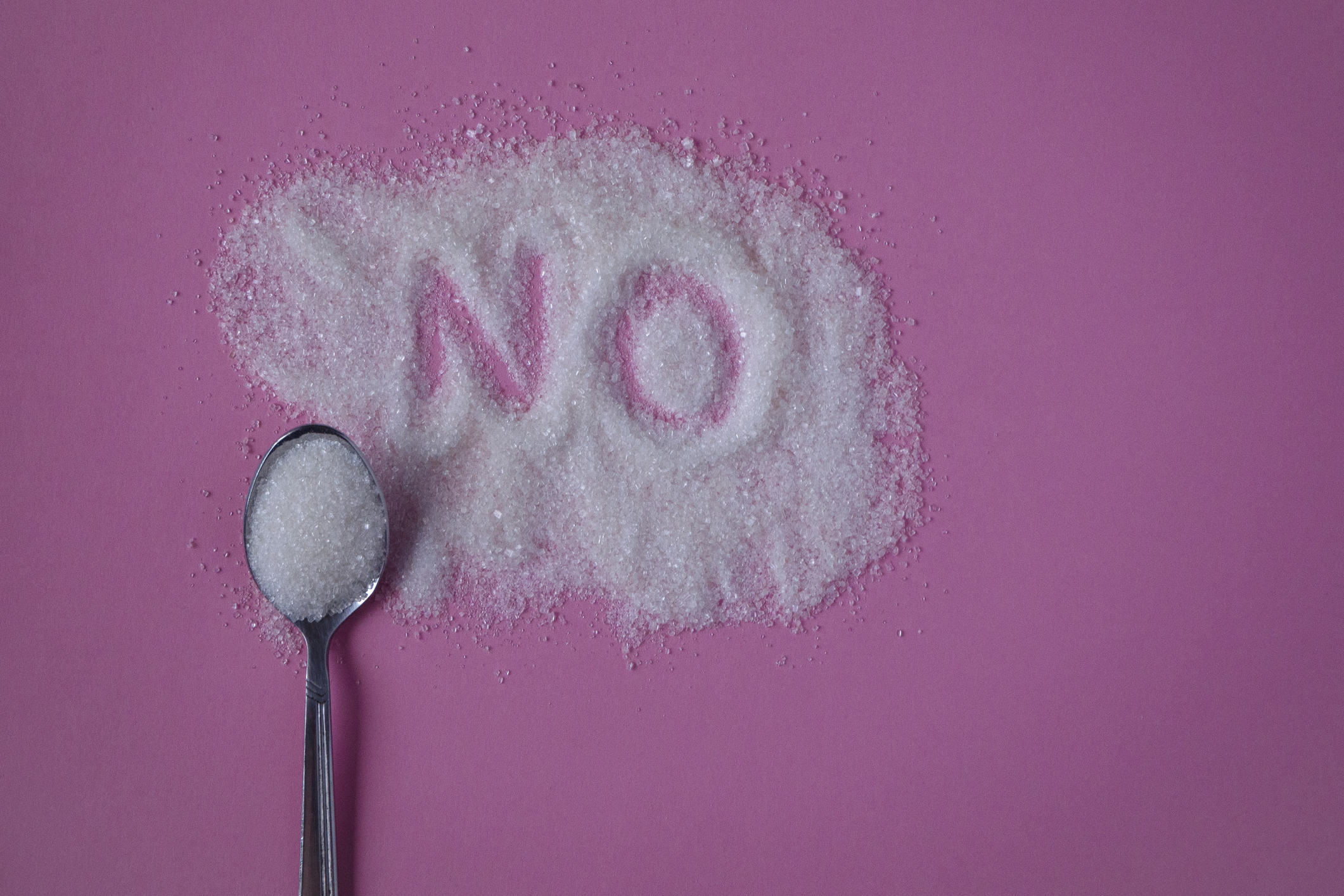 Word NO hand written on a heap of white sugar with full teaspoon of sugar on a pink background top view.