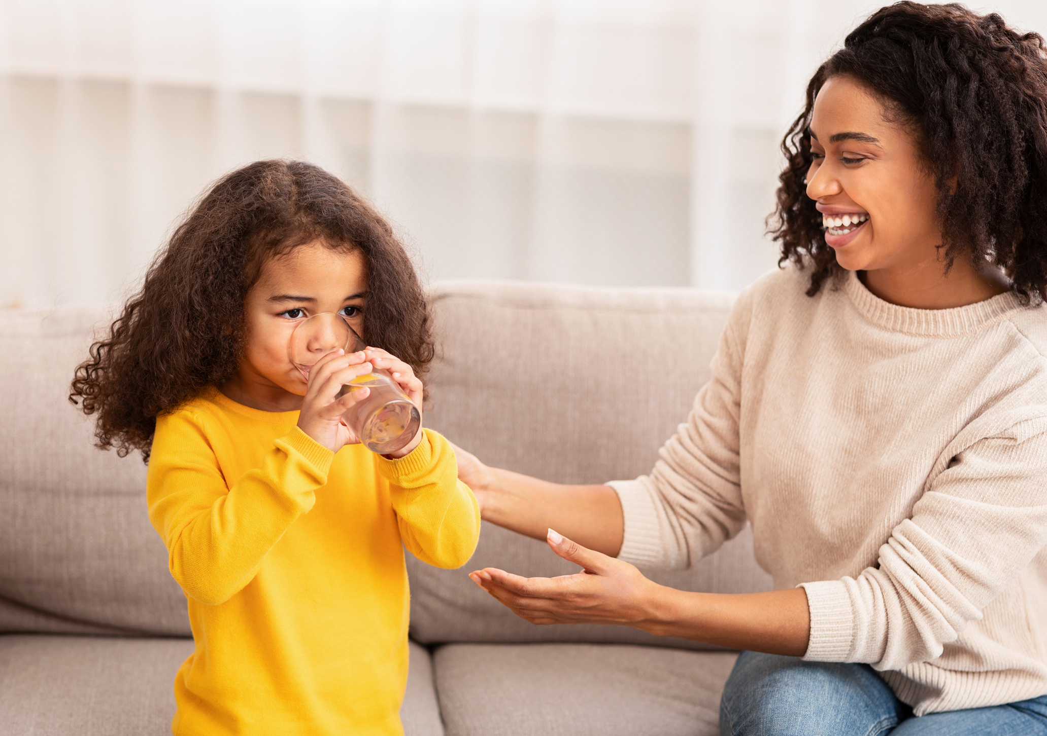 Woman Giving Daughter Glass Of Water Sitting On Sofa Indoor