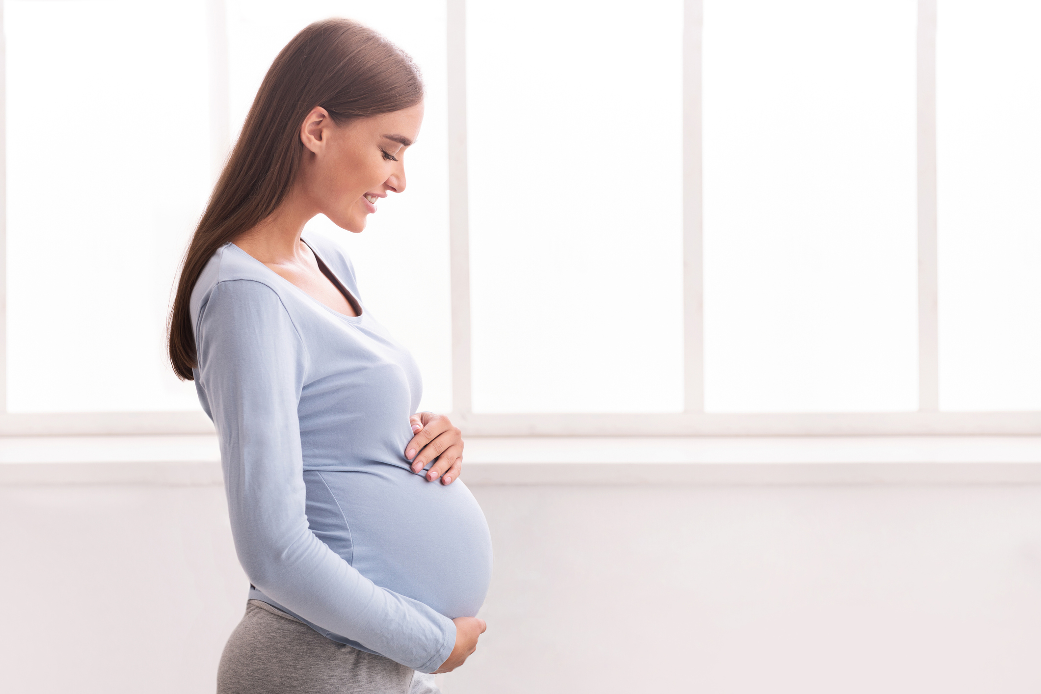 Joyful Expectant Woman Touching Belly Standing Near Window At Home