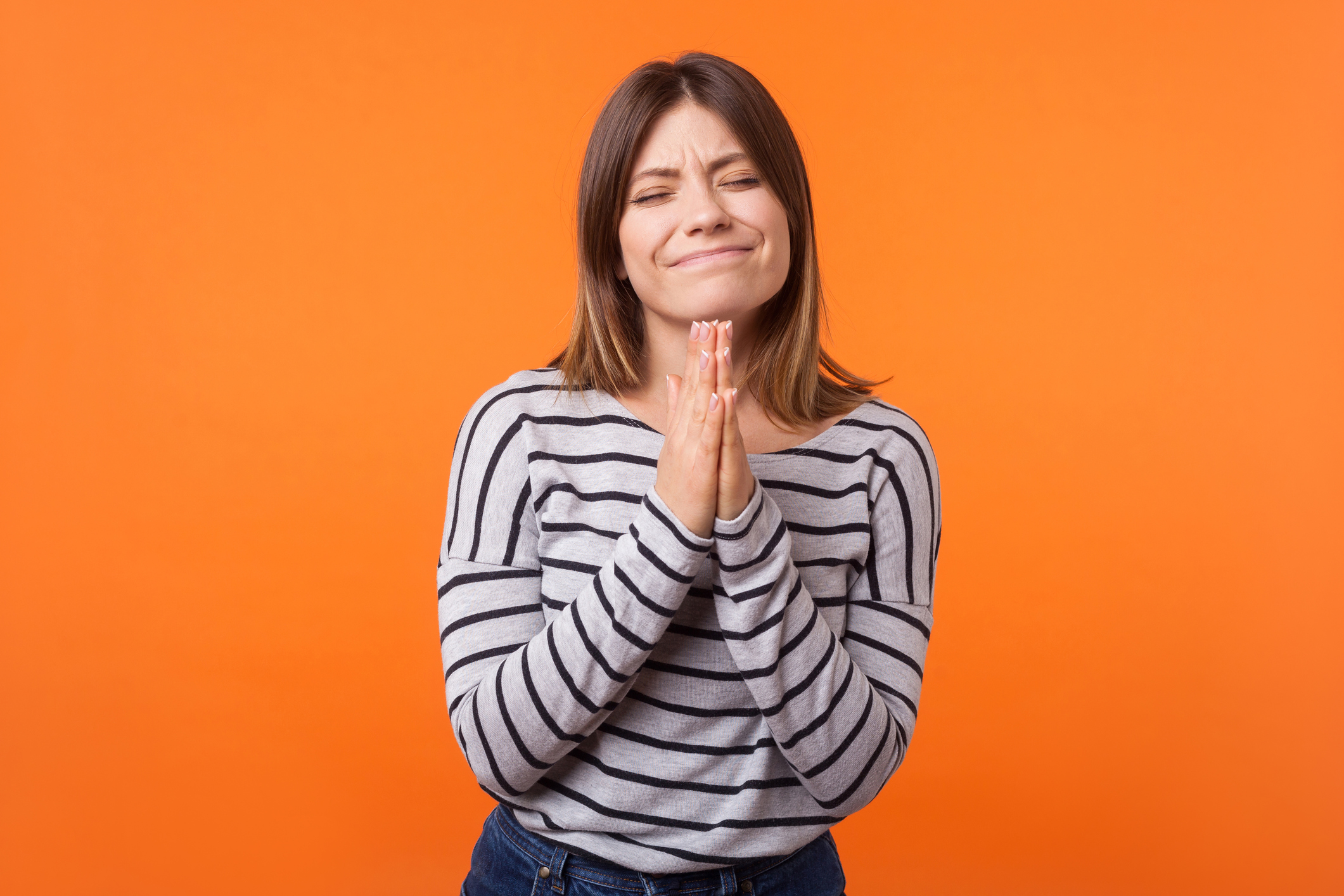 Please, let it happen! Portrait of hopeful young woman with brown hair in long sleeve striped shirt. indoor studio shot isolated on orange background