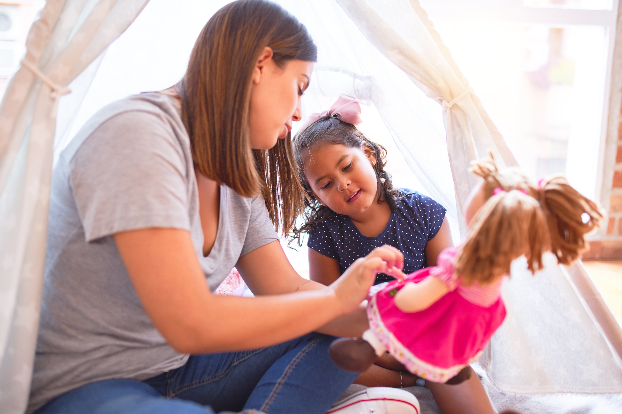 Beautiful teacher and toddler girl sitting on the floor playing with unicorn and doll inside tipi at kindergarten