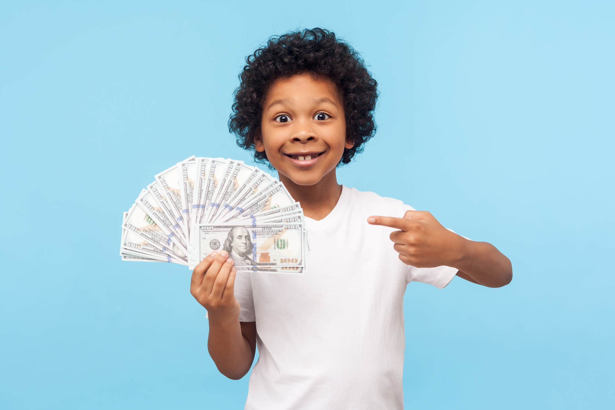 I'm rich! Joyful lucky wealthy little boy with curly hair pointing to fan of dollar banknotes