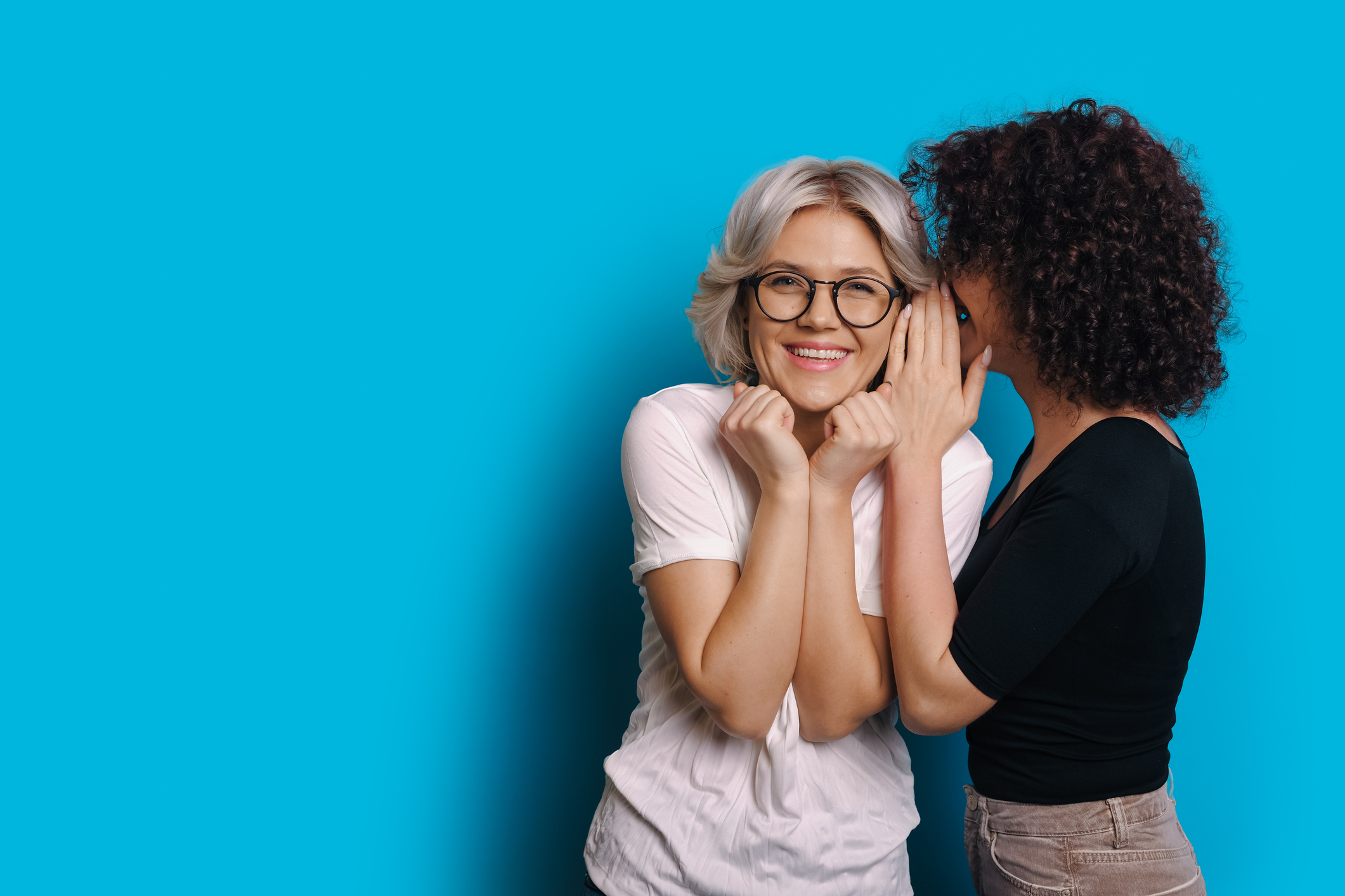 Curly haired caucasian girl is talking something secret to her blonde friend who is wearing eyeglasses and posing on a blue blank space background