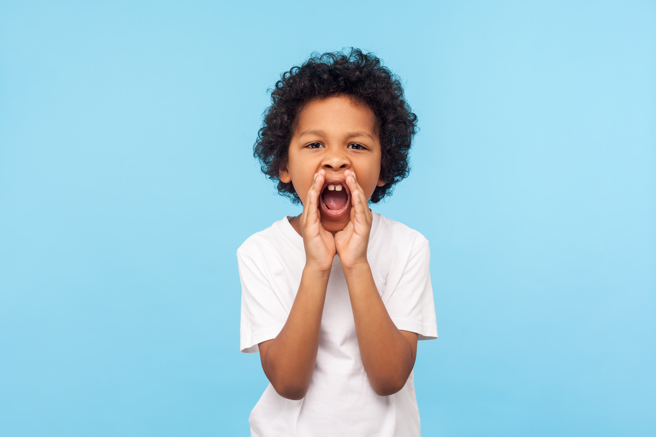 Attention! Portrait of little boy with curly hair in white T-shirt holding hands near wide open mouth and shouting loudly