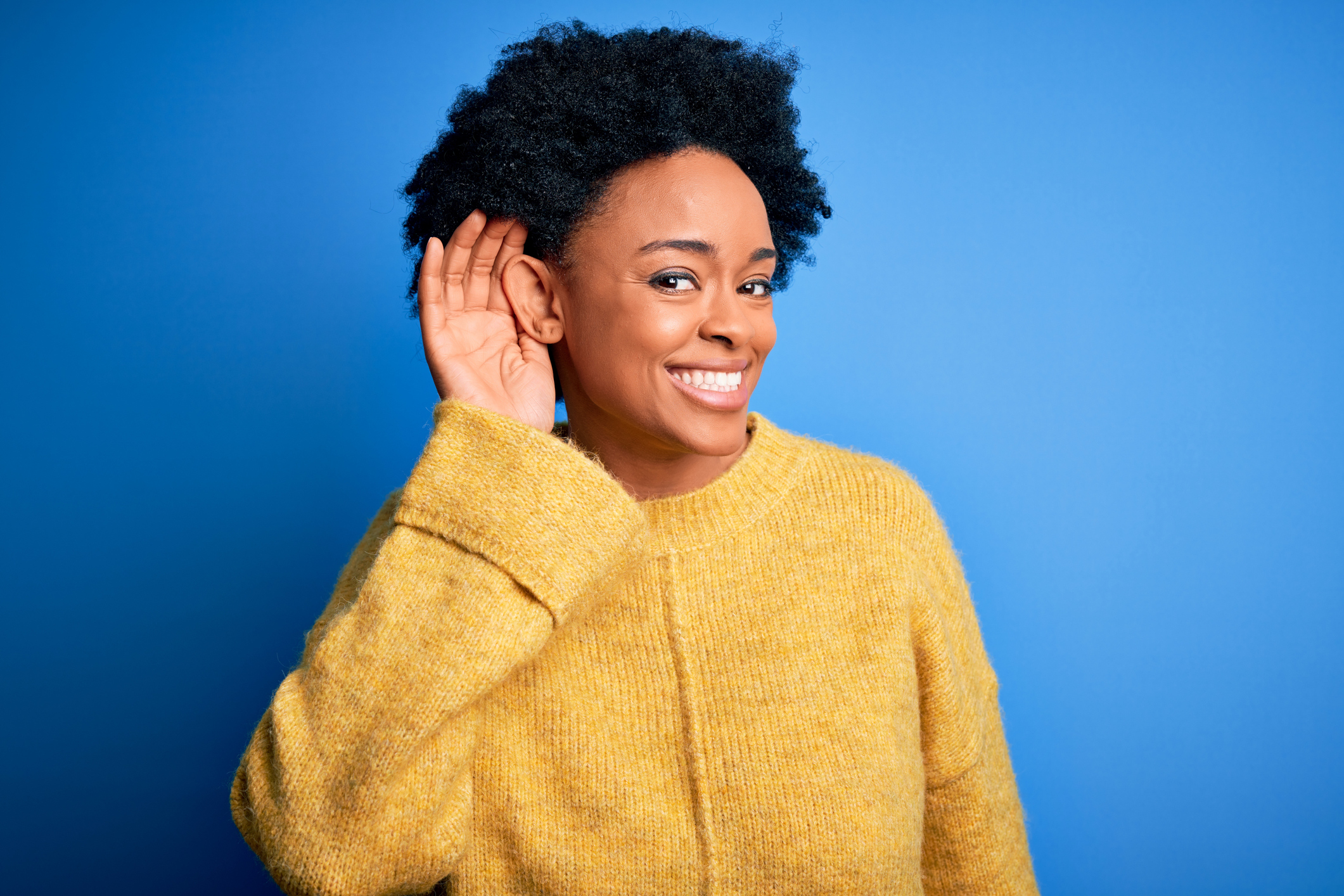 Young beautiful African American afro woman with curly hair wearing yellow casual sweater smiling with hand over ear listening an hearing to rumor or gossip. Deafness concept.