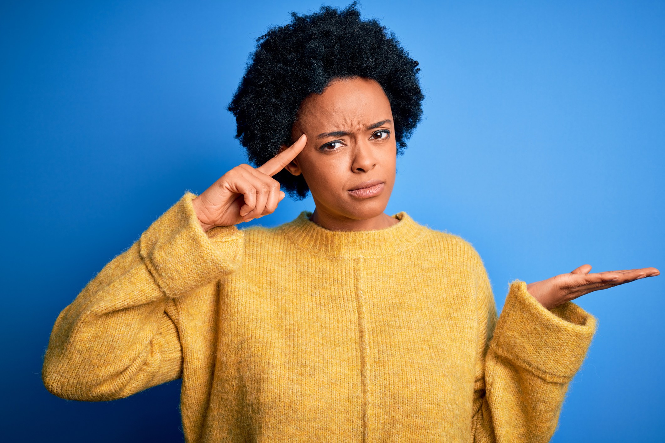 Young beautiful African American afro woman with curly hair wearing yellow casual sweater confused and annoyed with open palm showing copy space and pointing finger to forehead. Think about it.