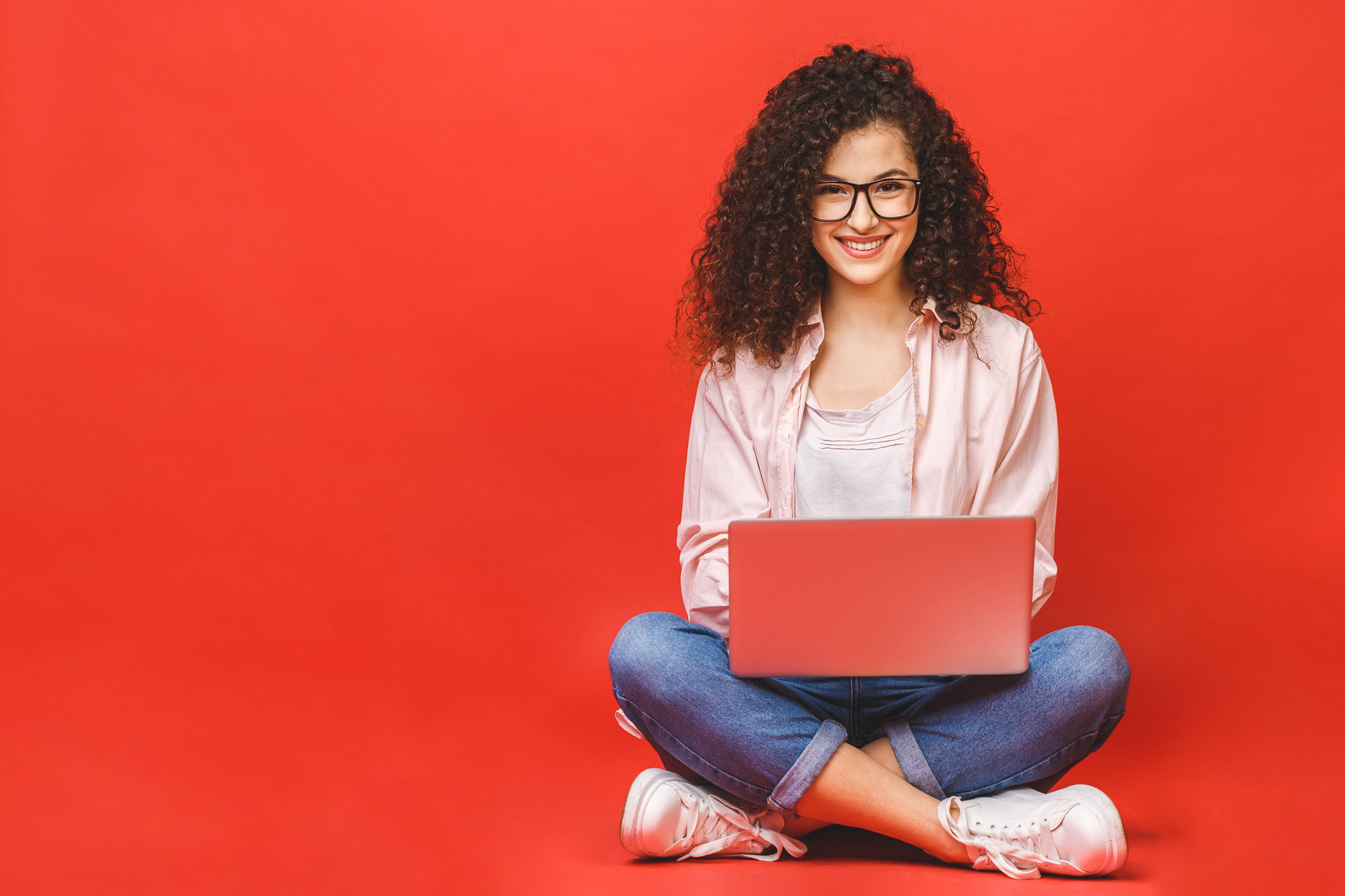 Happy young curly beautiful woman sitting on the floor with crossed legs and using laptop on red background.