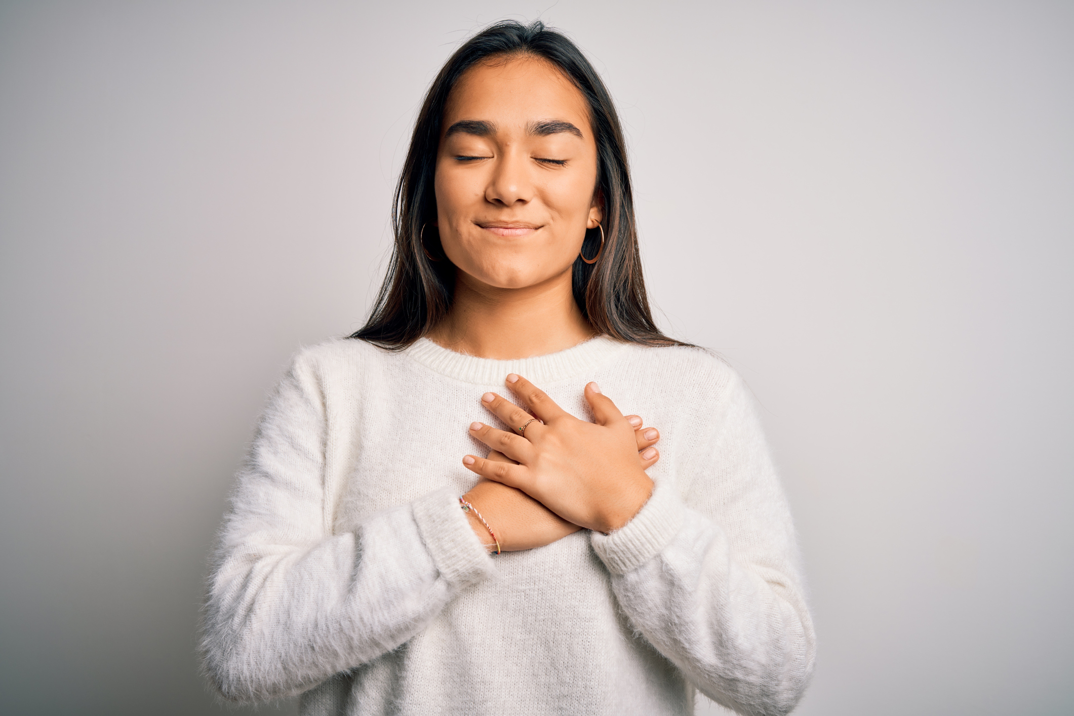 Young beautiful asian woman wearing casual sweater standing over white background smiling with hands on chest with closed eyes and grateful gesture on face. Health concept.