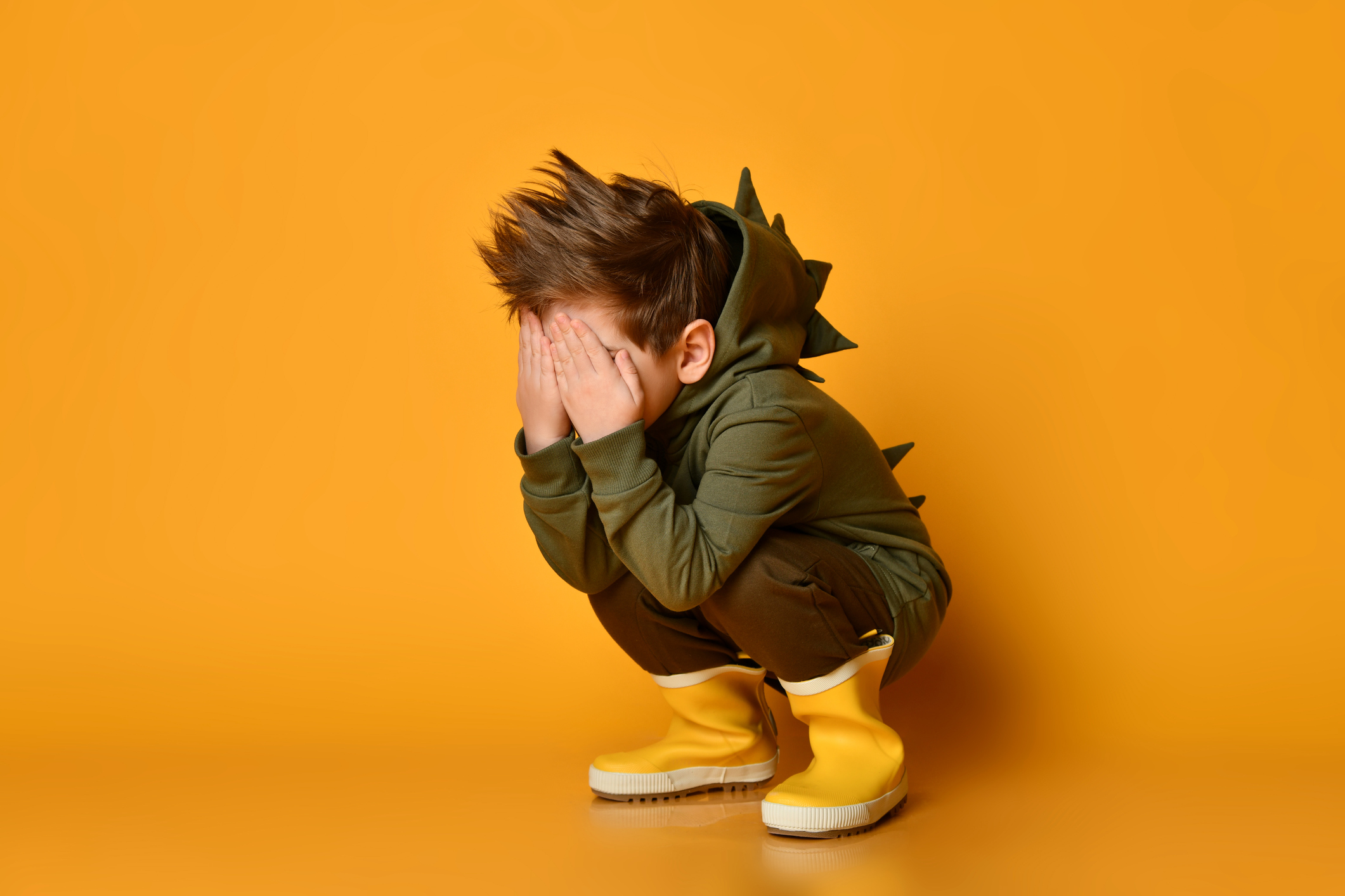 Kid boy in hoodie with dino spikes at back and cowl and in yellow rubber boots sits squatting covering face with hands