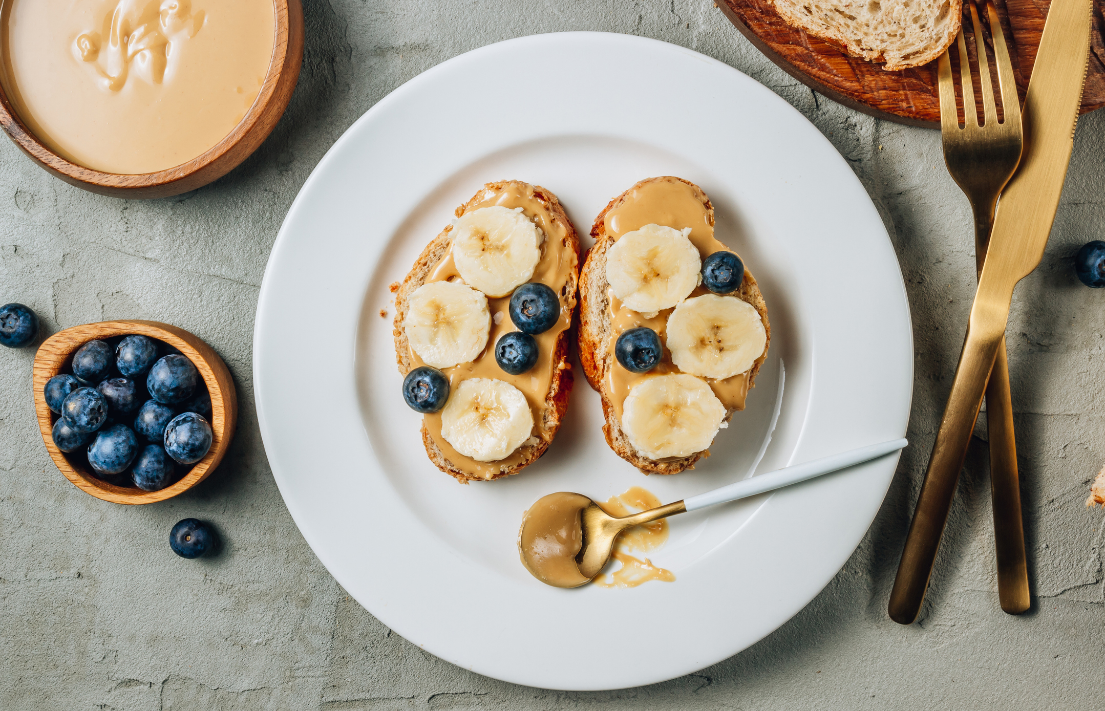 Buckwheat healthy bread with peanut butter, banana and blueberry on white plate over concrete background. Top view. Flat lay. Summer breakfast.