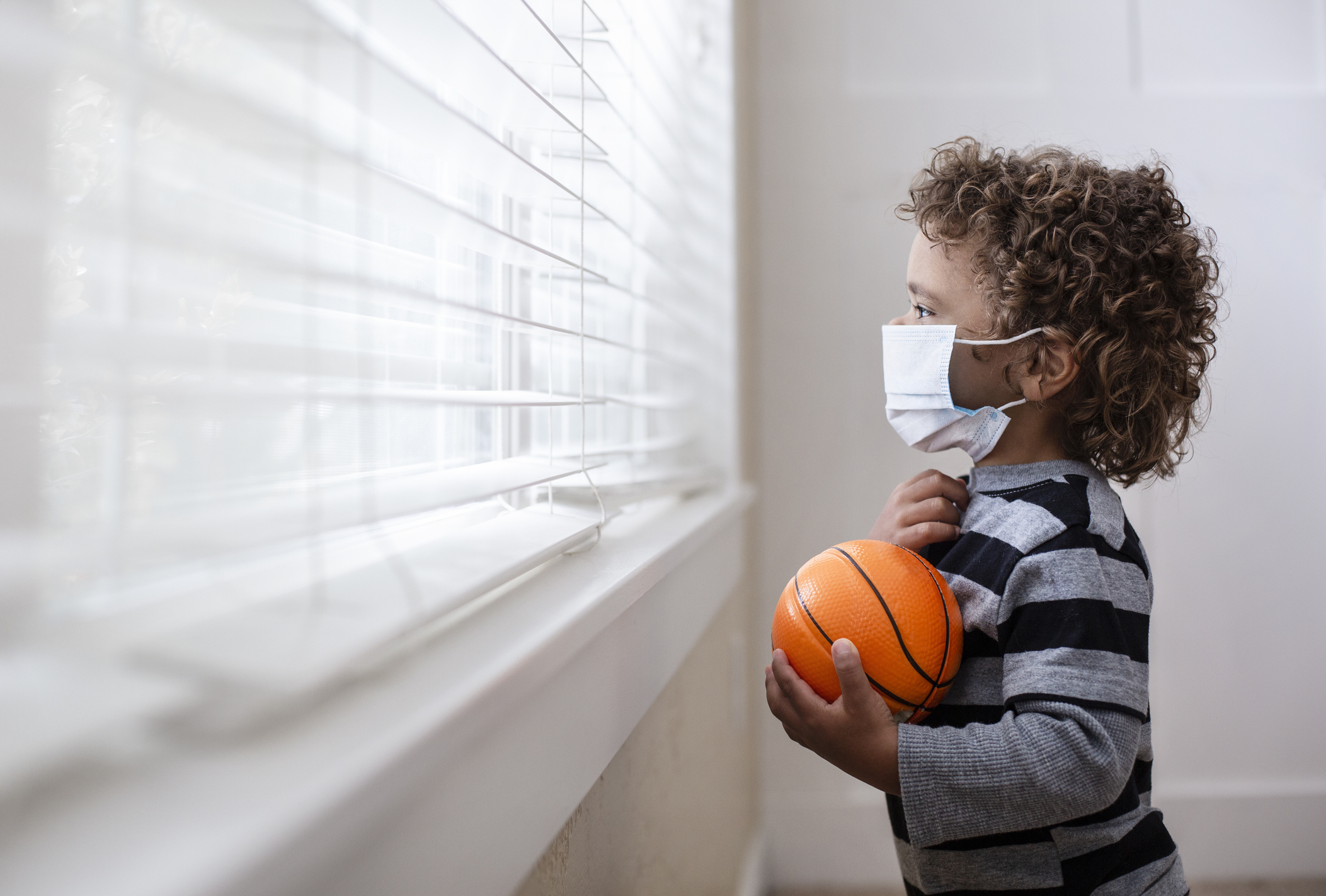 A young boy looking out the window wearing a protective facemark while seeking protection from COVID-19