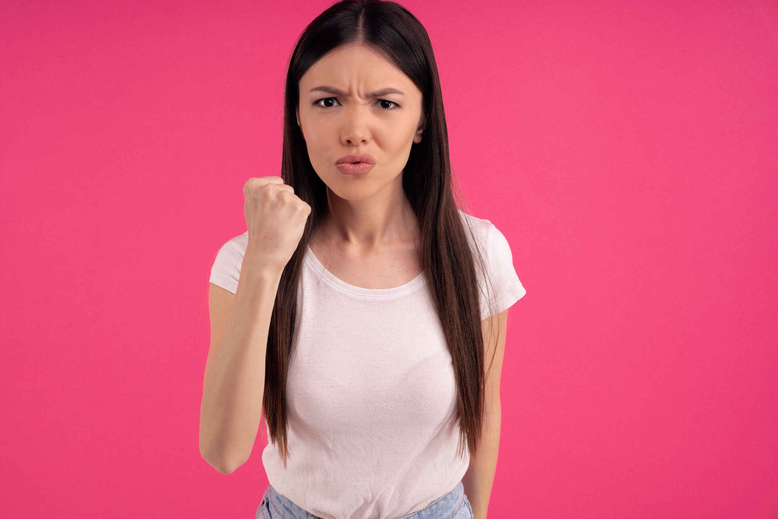 Picture of angry dissatisfied Asian woman clenches fist with displeasure, keeps lips folded, makes outraged face expression, wears white clothing, threatens you, isolated on pink background.