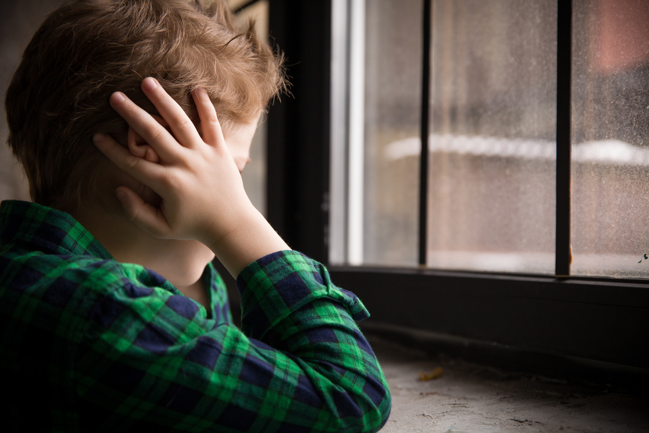 Little boy standing behind the window in sad mood. Sad Teenager looking in the Window and closing his ears with hands. Unhappy child in a plaid shirt. Alone at home. Upset.