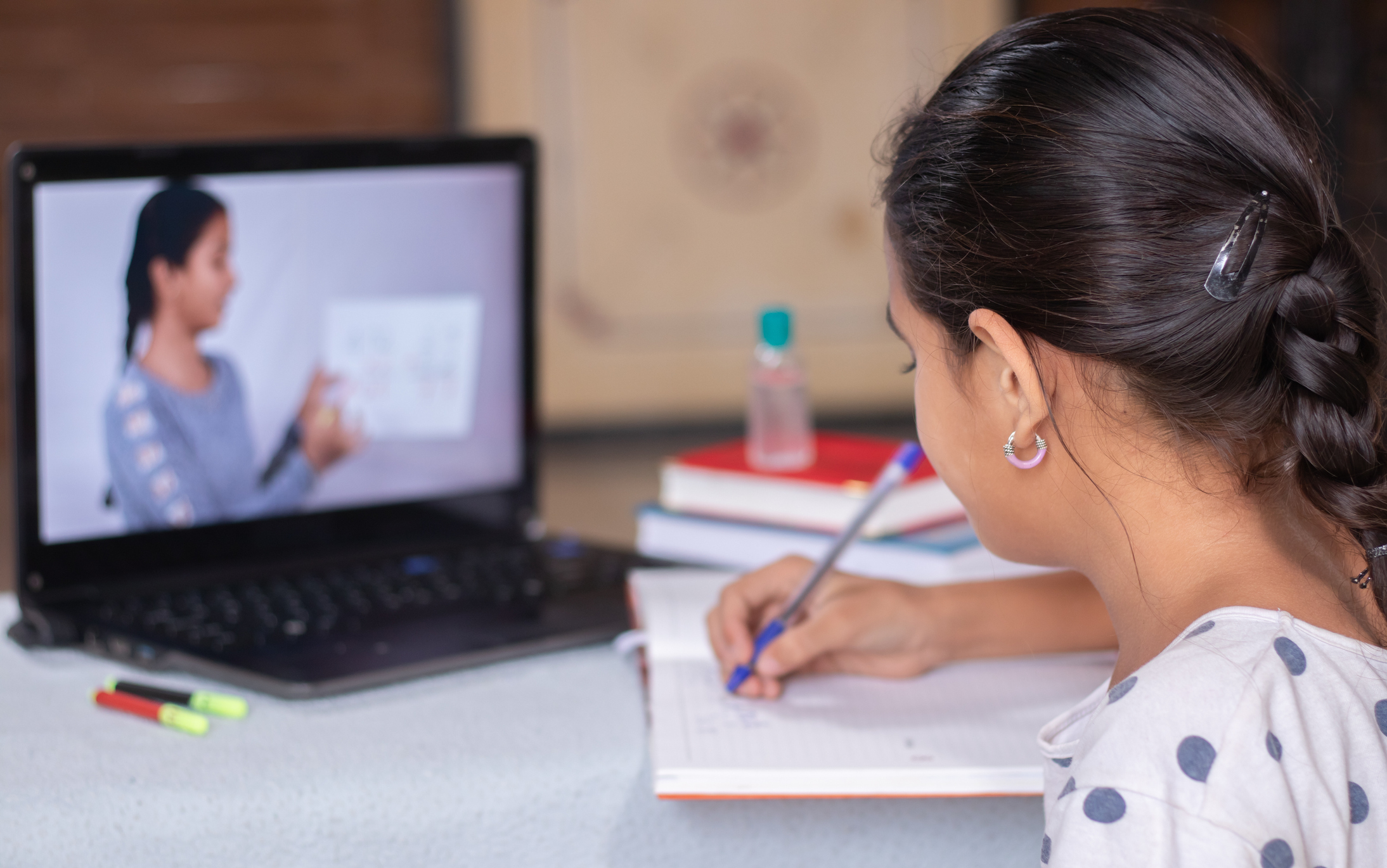 Concept of homeschooling or e-learning, young girl busy in writing by looking into laptop while teacher explaining during covid-19 or coronavirus pandemic crisis.