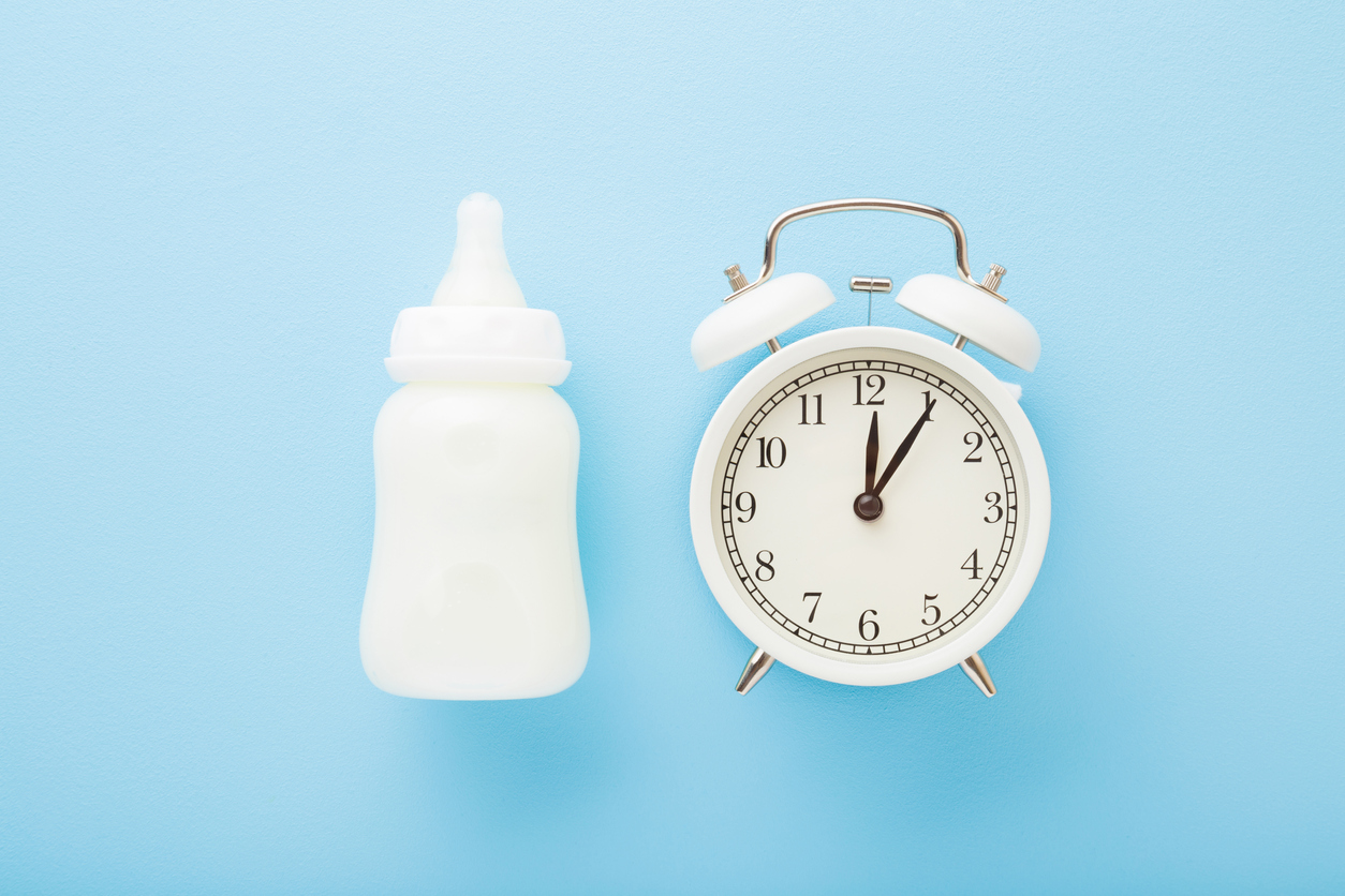 Alarm clock and bottle of white milk on light blue table background. Pastel color. Closeup. Baby feeding time. Top down view.