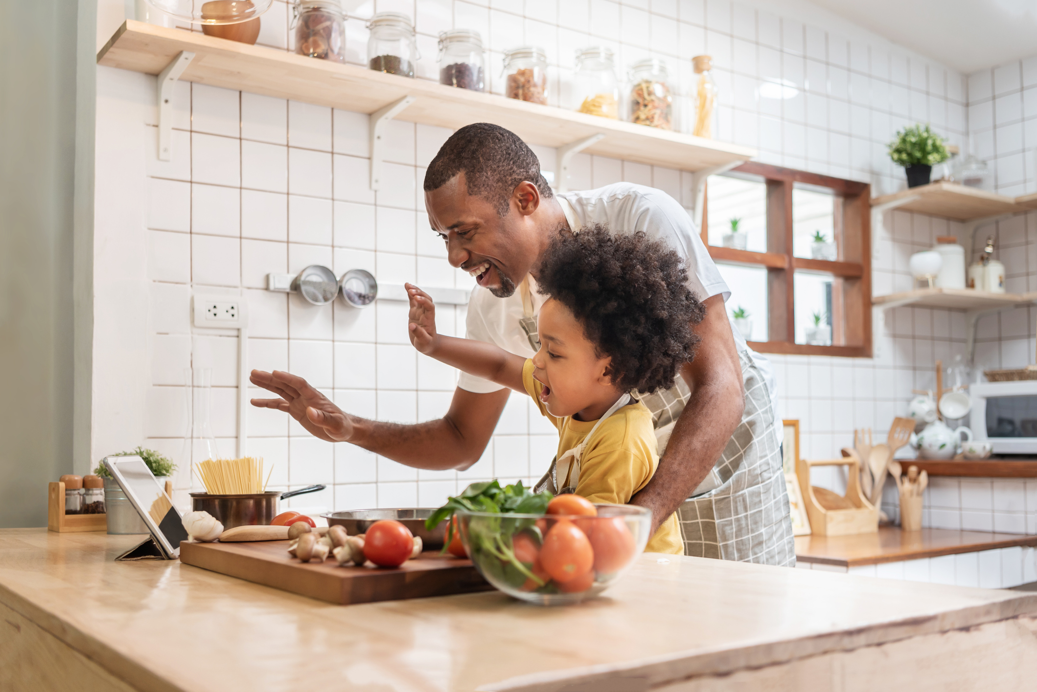 African American Father and little son making video call with digital tablet and waving hands laughing talking with family while cooking in kitchen. Black family have fun while pandemic virus at home