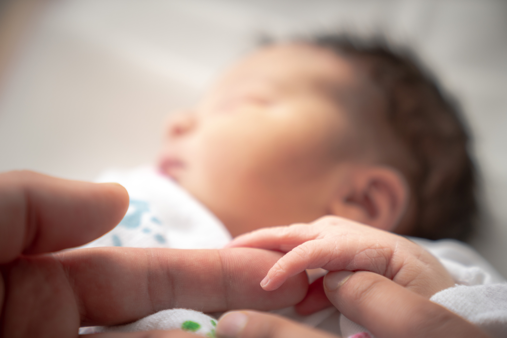 A newborn infant baby girl in a blanket swaddle wraps her tiny hand and fingers around her father and mother's fingers as she sleeps peacefully.