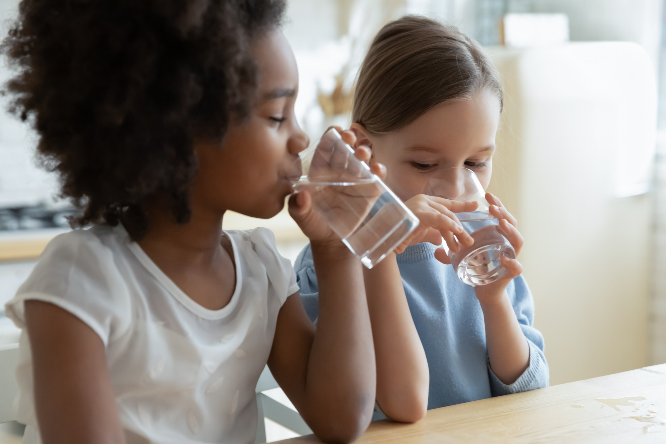 Two multiracial girls sit in kitchen feels thirsty drink water