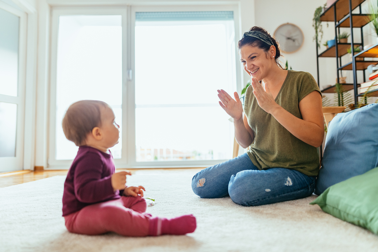 Nanny playing with baby girl at home - woman showing how to clap to baby