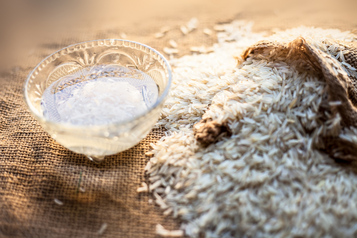 Shot of rice water in glass bowl with gunny bag full of basmati white rice in it over a brown surface.