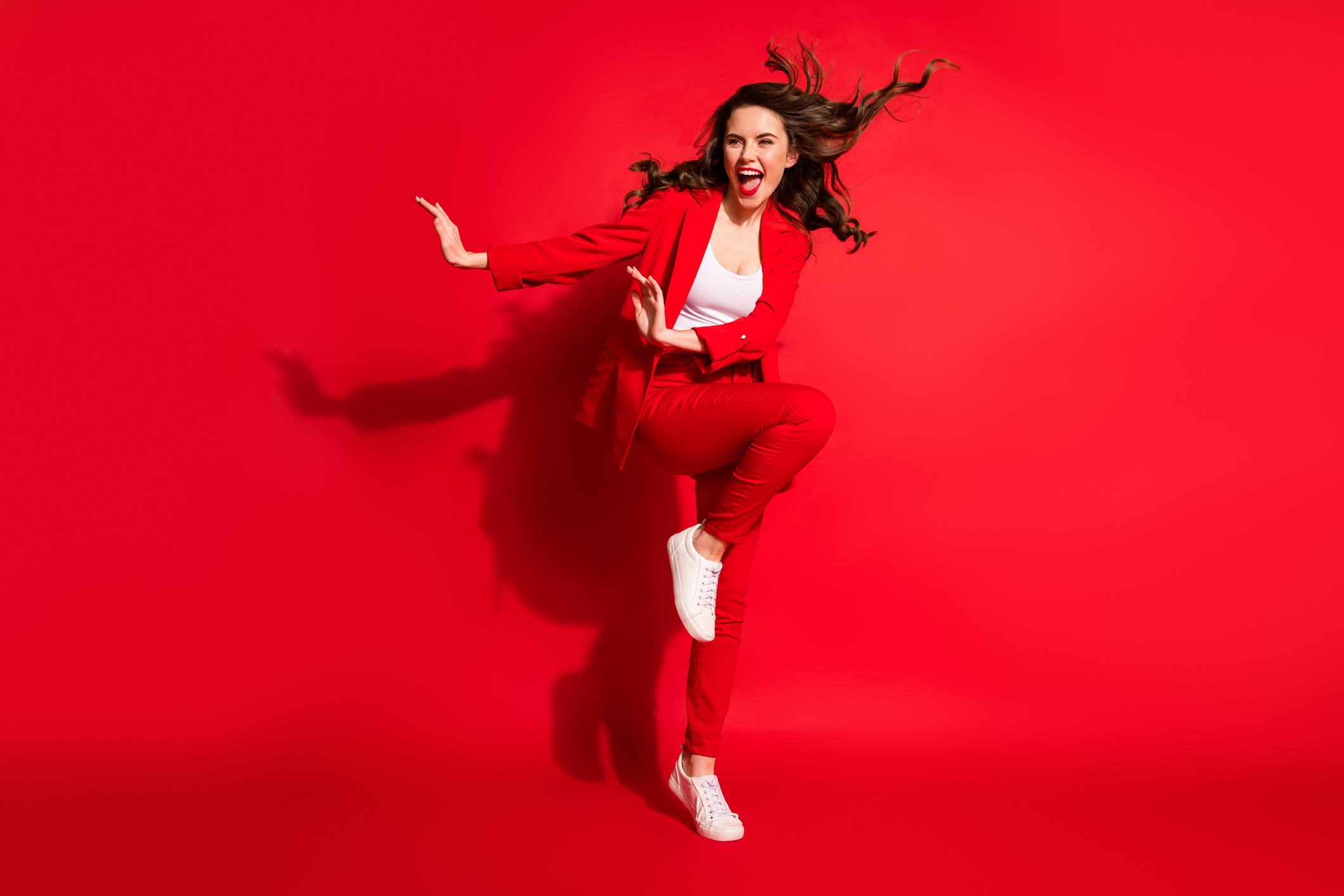 Full length photo of lovely young girl wind blow hairdo raise knee look side empty space wear jacket pants footwear isolated red color background