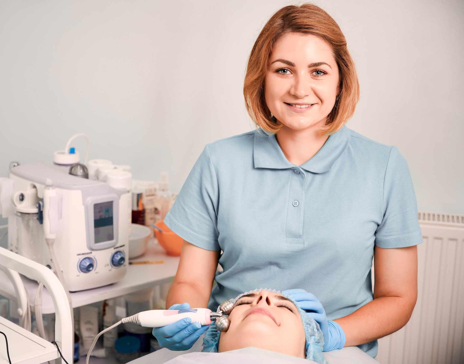 Cheerful beautician massaging woman face with facial roller.