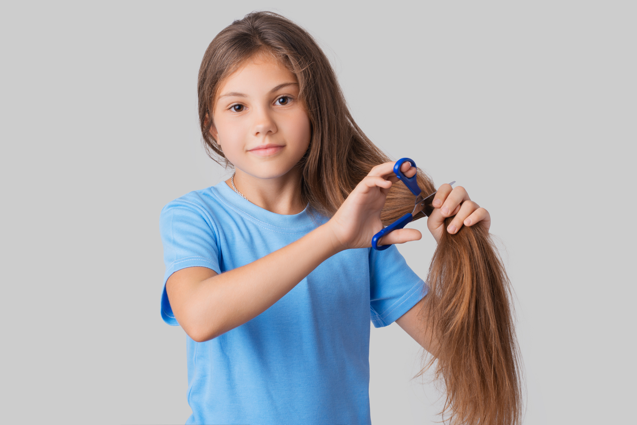 Small apple-cheeked girl in a blue t-shirt going to cut off her long hair with blue scissors against a grey background