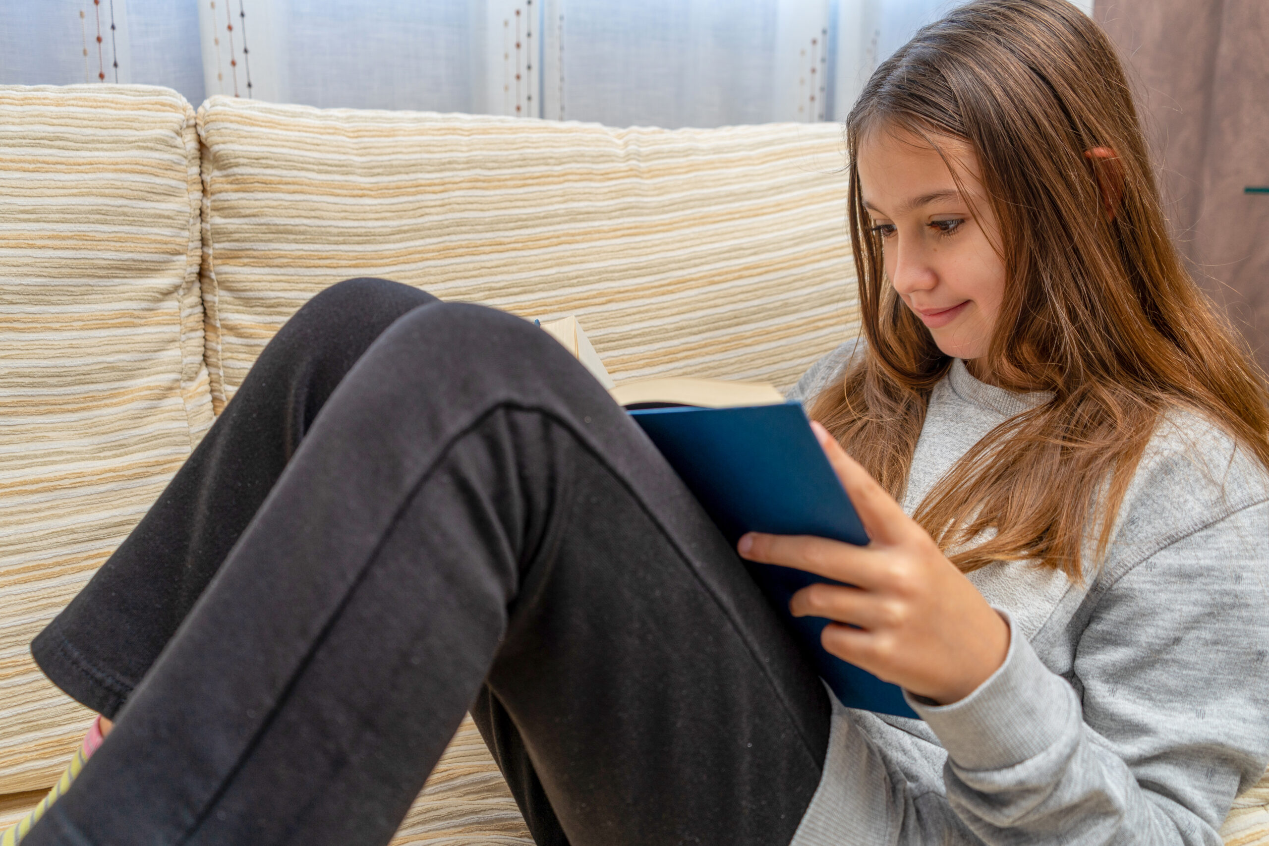 Cute little girl reading a book with interest on the sofa in her living room. Selective focus. Learning concept.