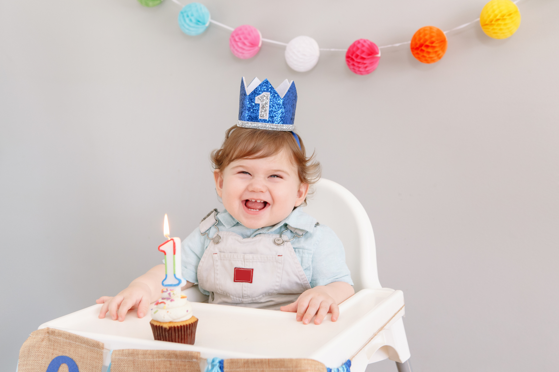 Happy smiling cute Caucasian baby boy in blue crown celebrating his first birthday at home. Child kid toddler sitting in high chair eating tasty cupcake dessert with a lit candle. Happy birthday concept