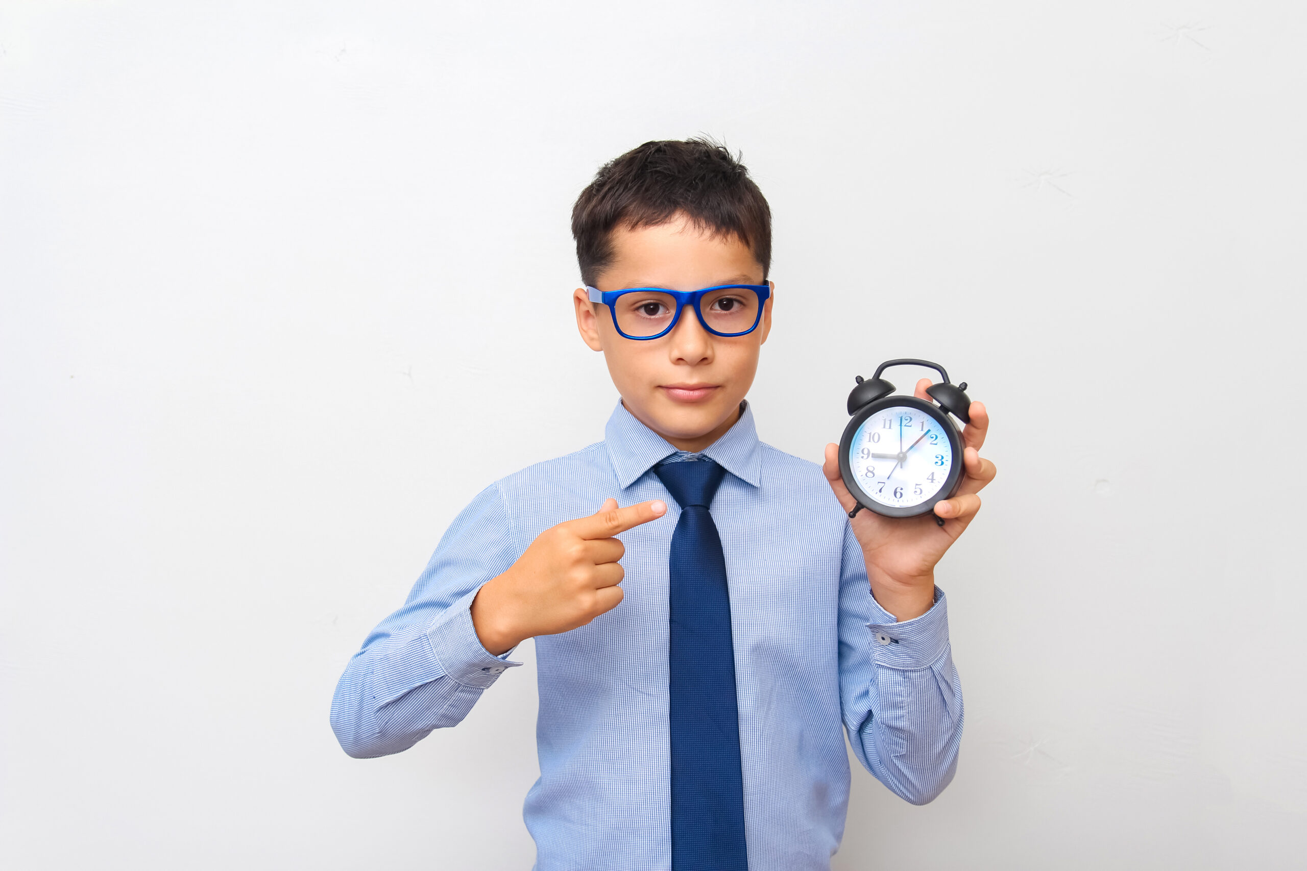 A dark-haired Caucasian boy in a blue shirt and glasses holds an alarm clock in his hand and points at it with his finger, on a gray background. The concept of deadlines, time management