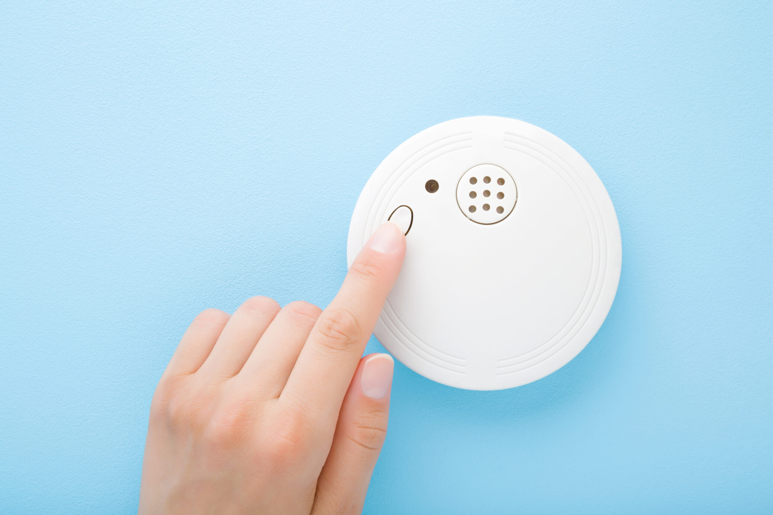 Young adult woman finger pushing button of new white plastic smoke alarm. Light blue table background. Pastel color. Closeup. Safety concept.