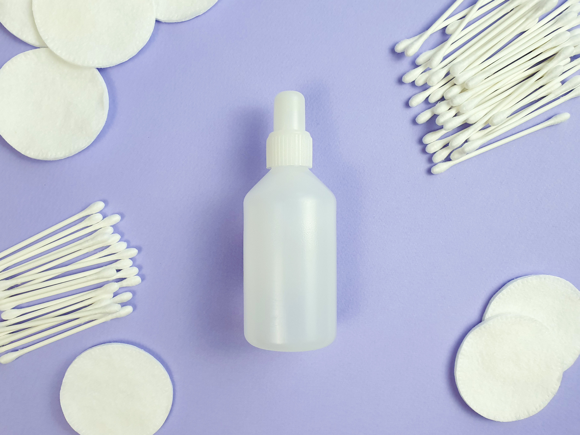 Unbranded white plastic bottle of disinfectant, cotton pads and sticks on purple background. Natural organic spa cosmetics and liquid antimicrobial concept. Flatlay style, top view.