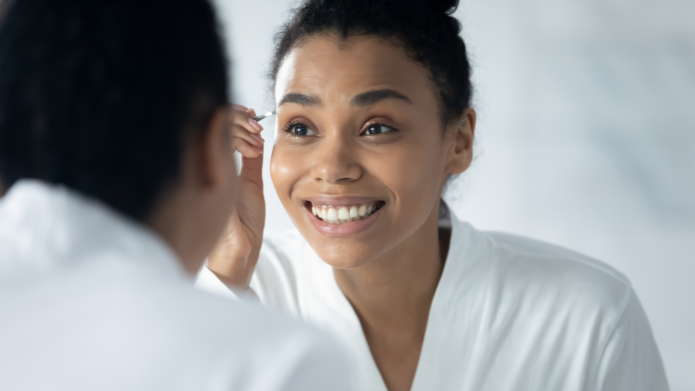 Happy engaged Black millennial girl in bathrobe plucking eyebrows