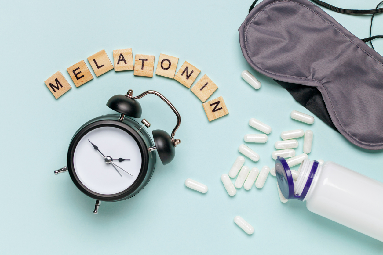 White bottle and pills, letters Melatonin,  alarm clock and sleep mask on light grey background. Sleeping backdrop Concept melatonin, insomnia and good night. Top view