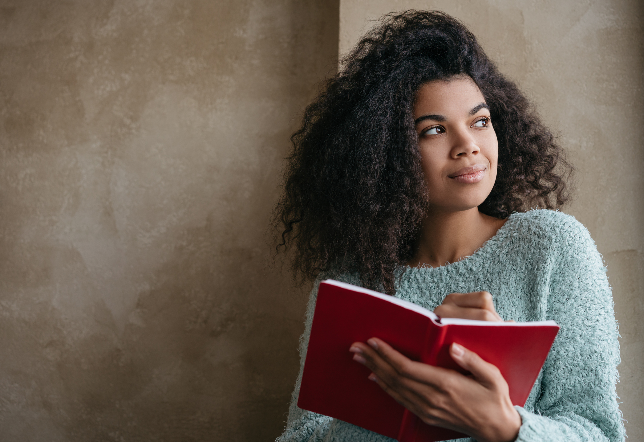 Beautiful African American woman holding red book, looking at window and smiling. University student studying, learning language, sitting at library. Portrait of young pensive writer taking notes