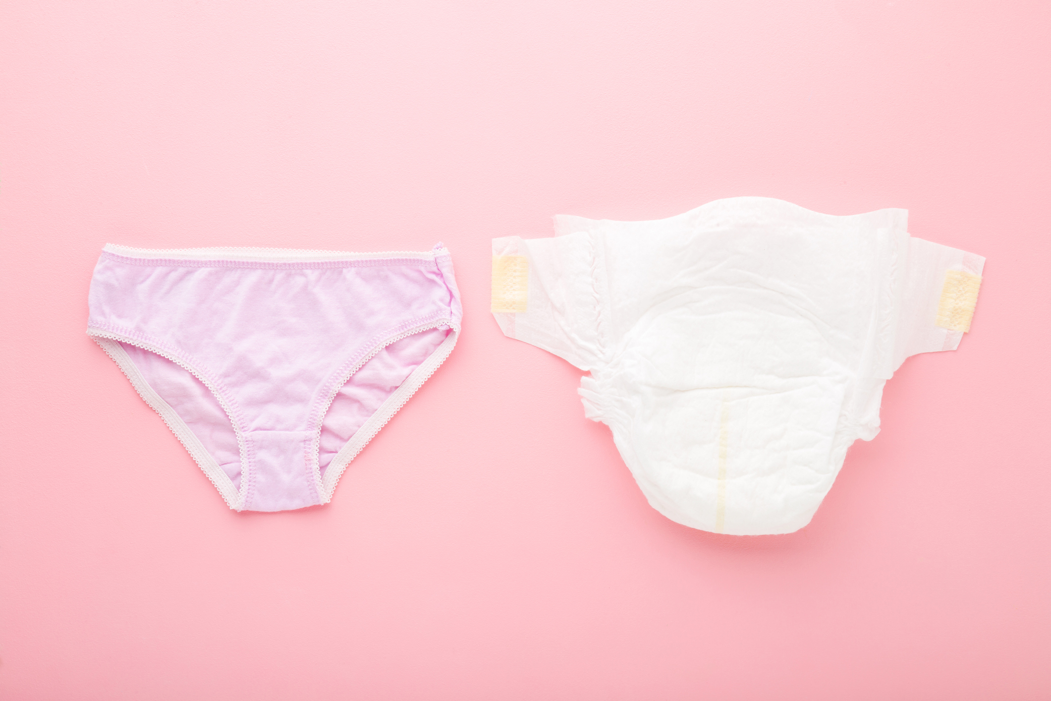 White soft nappy and light purple panties on pink table background. Pastel color. Closeup. Concept - when it is time to change baby diaper to underwear. Top down view.