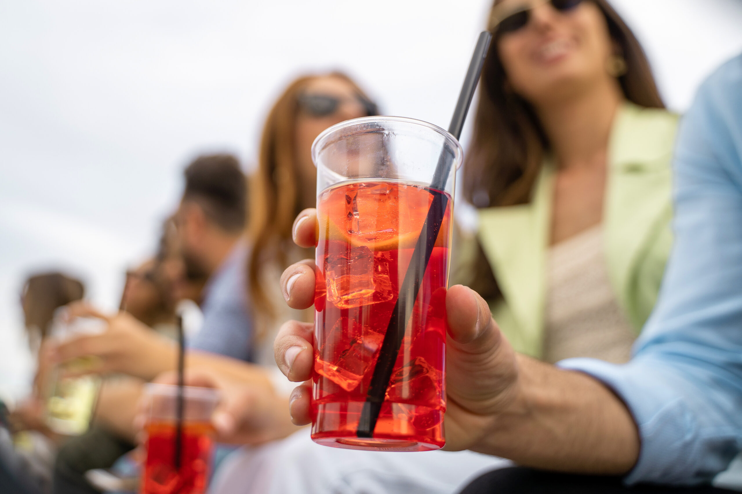 Gathering of best friends having fun talking and drinking cocktails together. Close up of an hand holding an iced spritz drink. Defocused young people on the background.