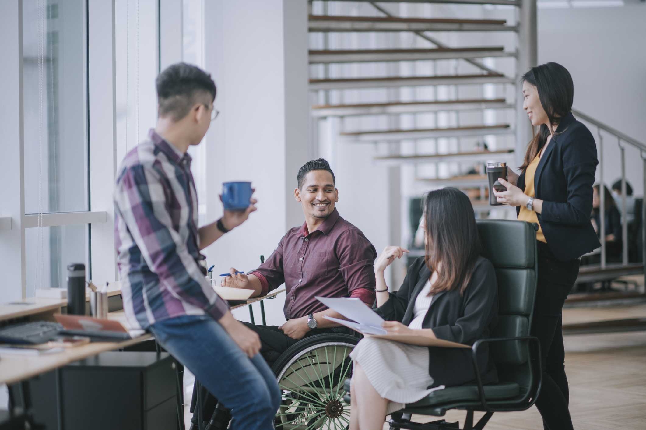 Indian white collar male worker in wheelchair having cheerful discussion leading conversation with colleague in creative office workstation beside window