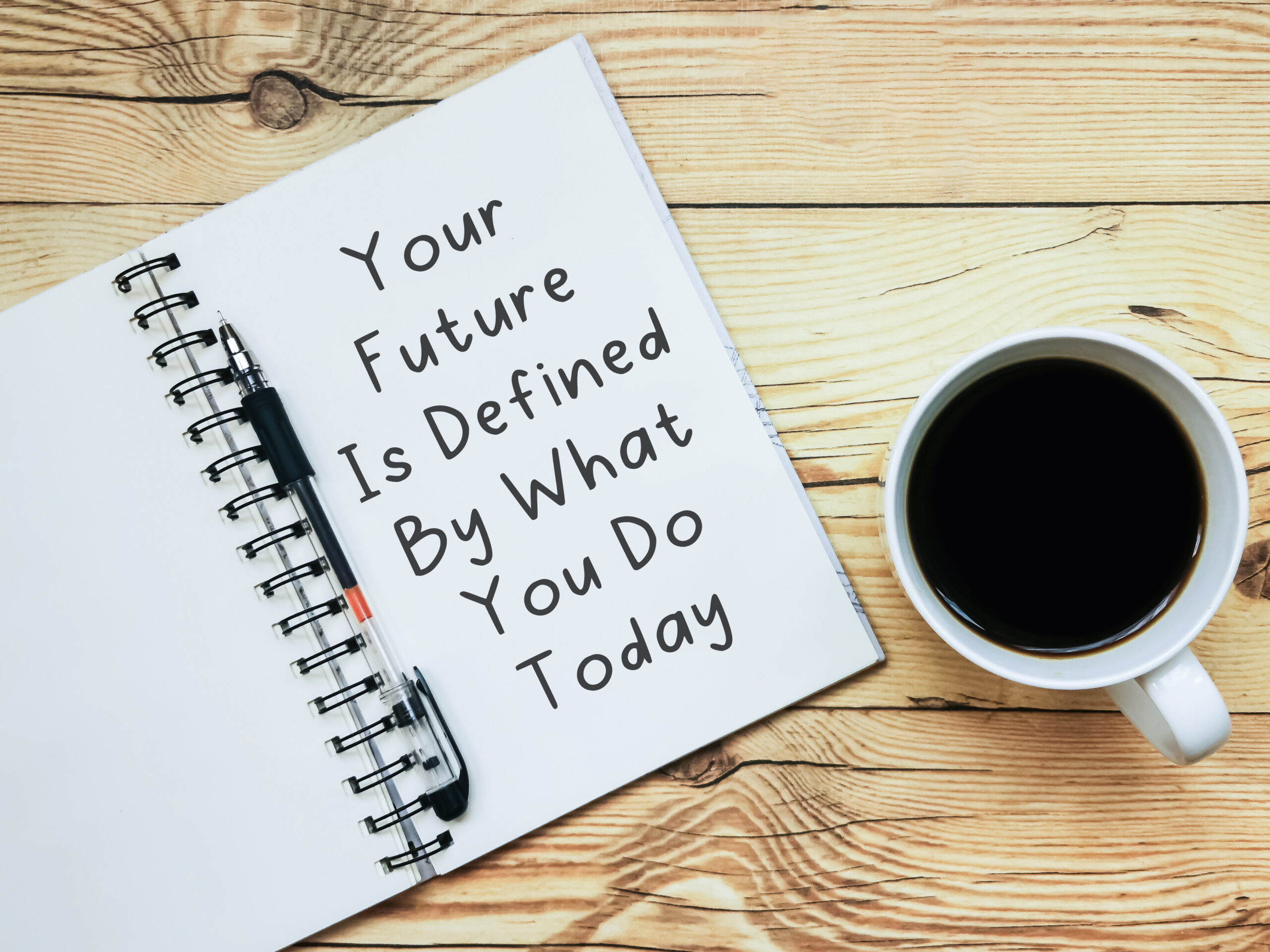 Open notebook with text "Your Future Is Defined By What You Do Today" and a cup  of coffee on wooden background.