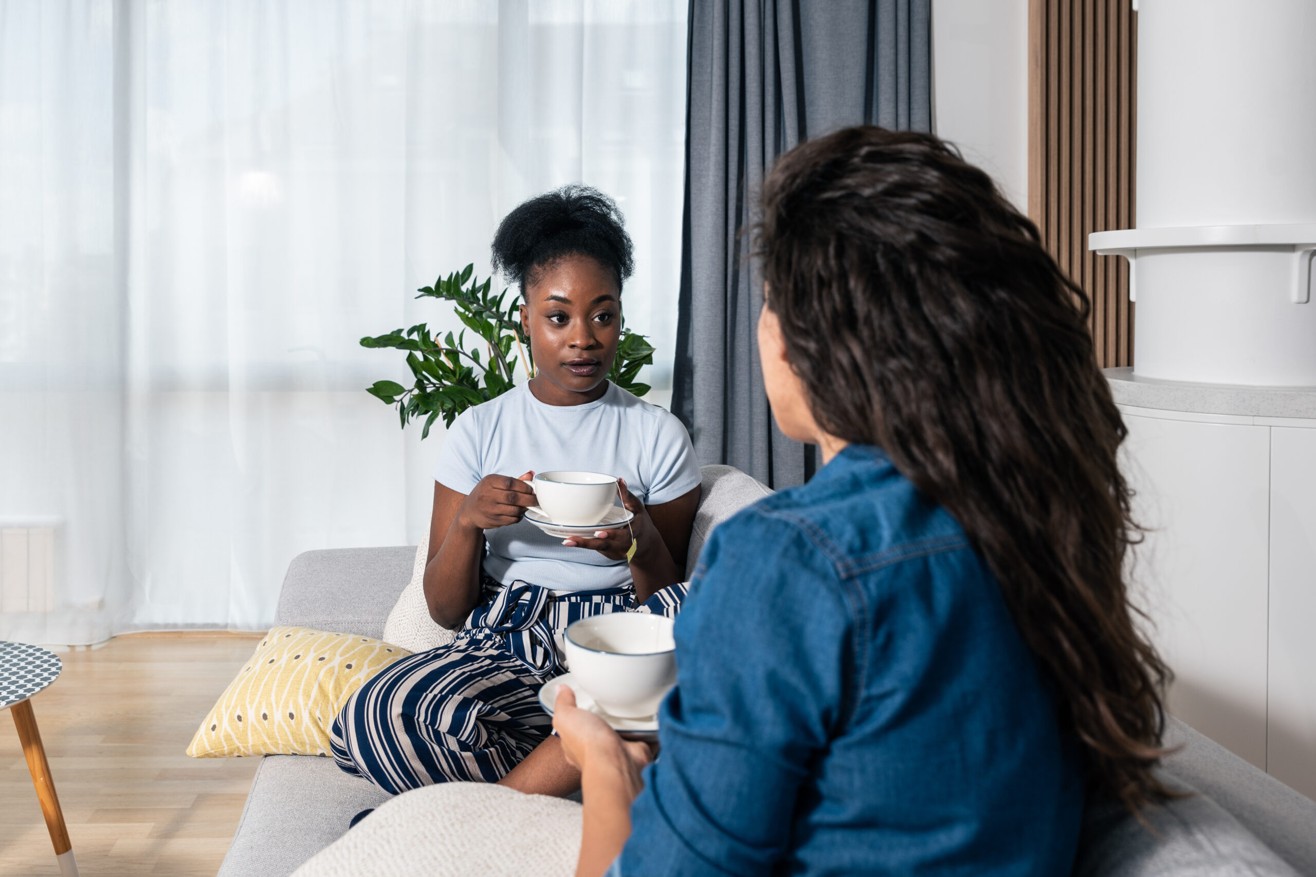 Two young women best friends sitting on the sofa at home drinking tea or coffee black African is complaining on her boyfriend or husband to white one how he changed and become jealous, selective focus