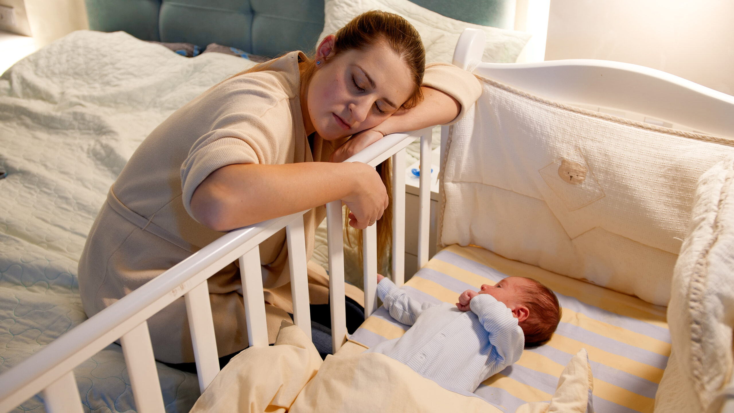 Young tired and exhausted mother fallen asleep while rocking crib of her newborn baby at night. Concept of sleepless nights and parent depression after childbirth