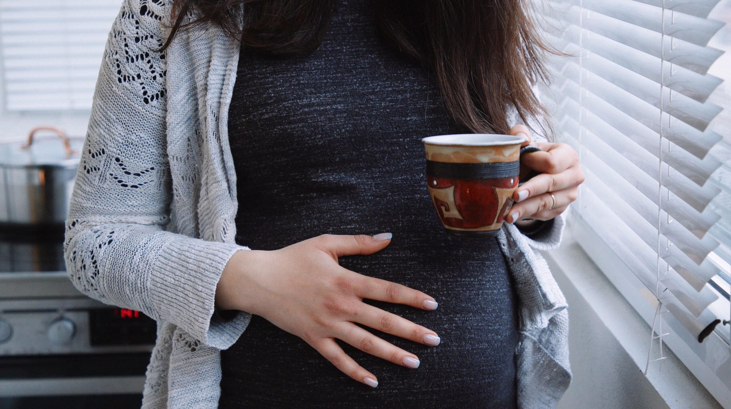 Pregnant woman caresses her belly while drinking  coffee next to the window at home.